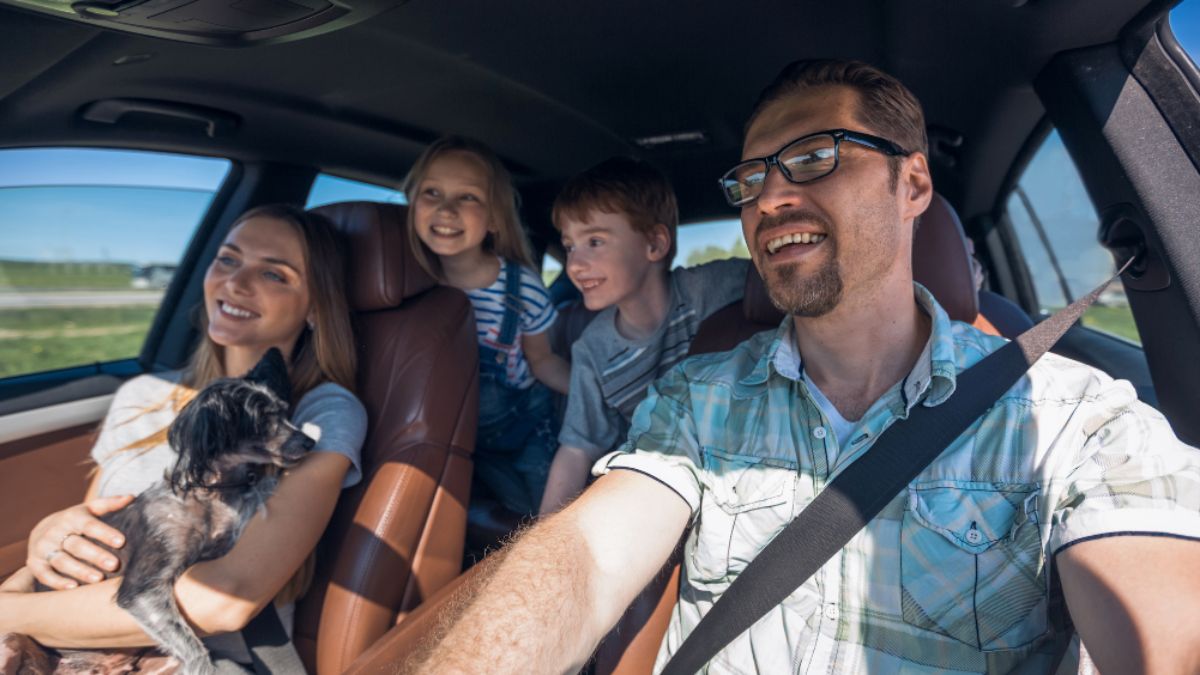 Smiling family and their dog enjoying a Family Road Trip, seen from the driver's perspective inside the car